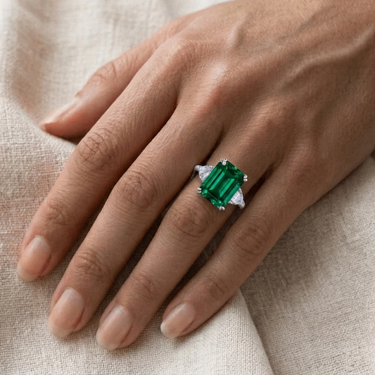 A close-up of a hand wearing a silver ring with a large emerald-cut green gemstone, resting on a textured beige fabric background.