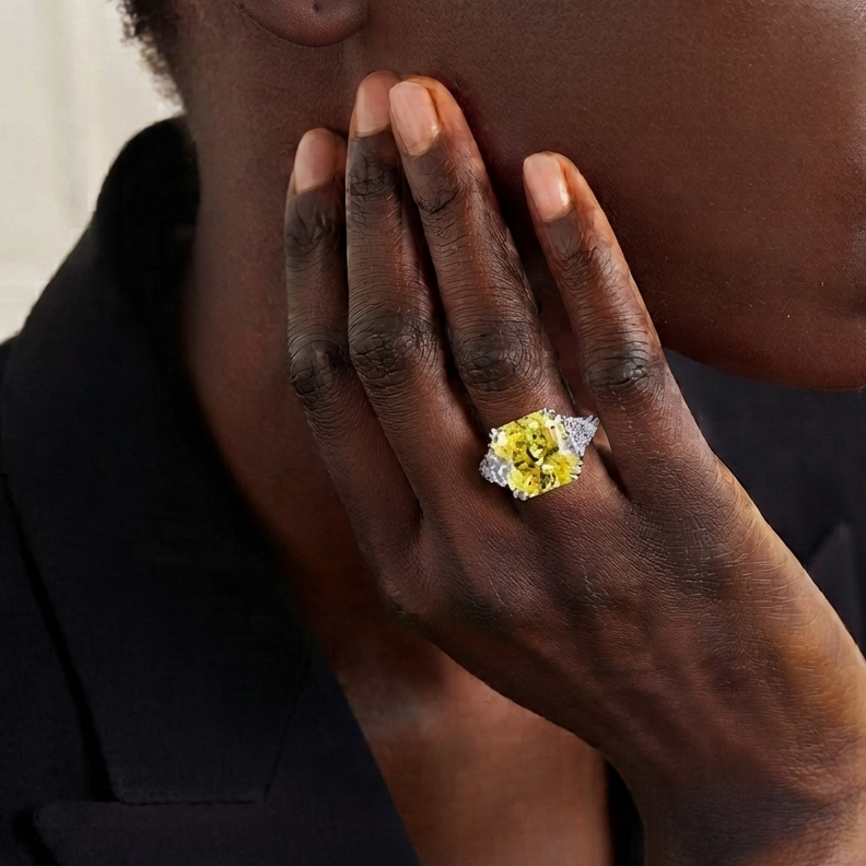 A close-up lifestyle shot of a model wearing a ring with a large yellow diamond on a silver band, set against a dark blazer and a neutral indoor background.