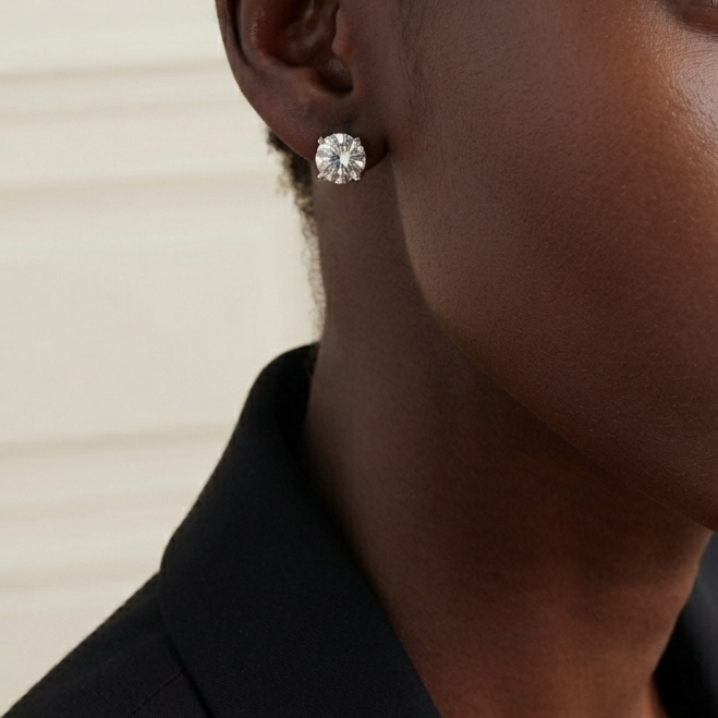 A close-up portrait of a model wearing a small, round brilliant-cut diamond stud earring. The model has short dark hair and is wearing a black top, set against a soft, light beige paneled background.